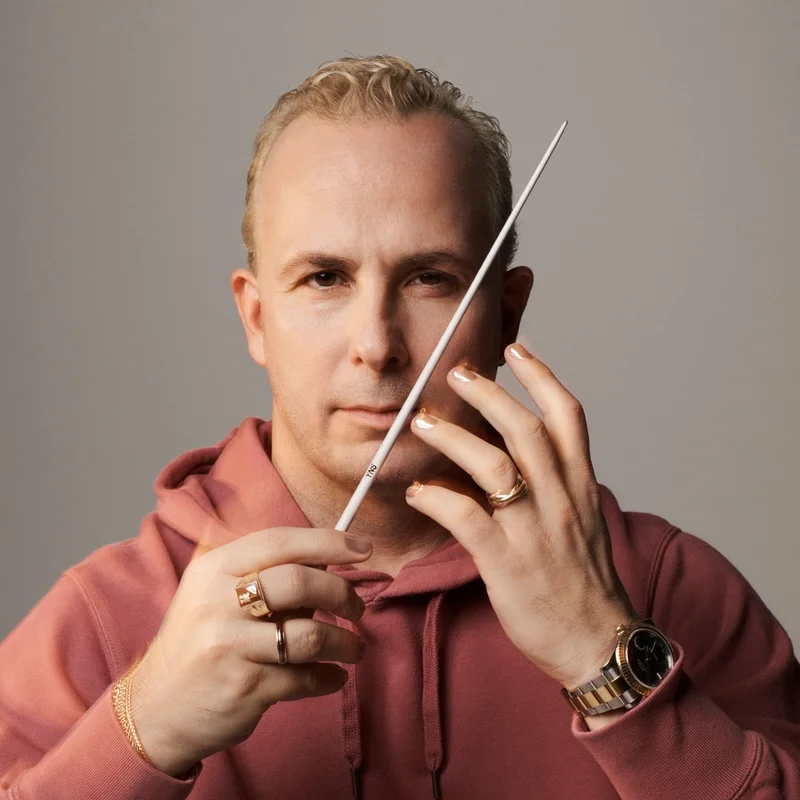 Yannick Nézet-Séguin in einem rosa Hoodie hält einen Dirigentenstab vor einem neutralen Hintergrund und hat einen konzentrierten Ausdruck.