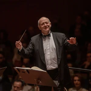 Ivan Fischer in formeller Kleidung leitet ein Orchester mit einem Taktstock, während das Publikum aufmerksam zuhört.
