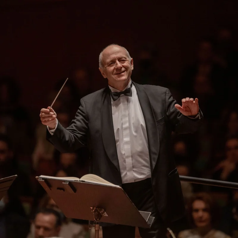 Ivan Fischer in formal attire leads an orchestra with a baton, while the audience listens attentively.