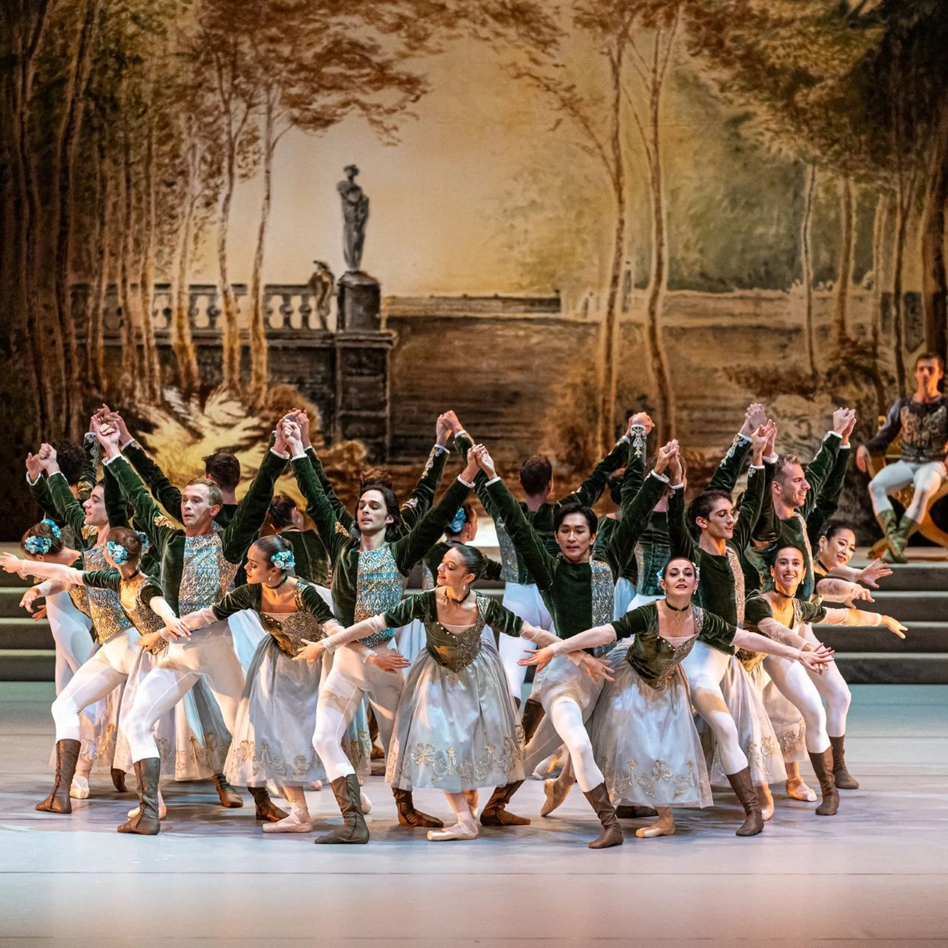 Eine Ballettgruppe führt vor einer malerischen Hintergrundkulisse auf. Die Tänzer tragen elegante historische Kostüme.