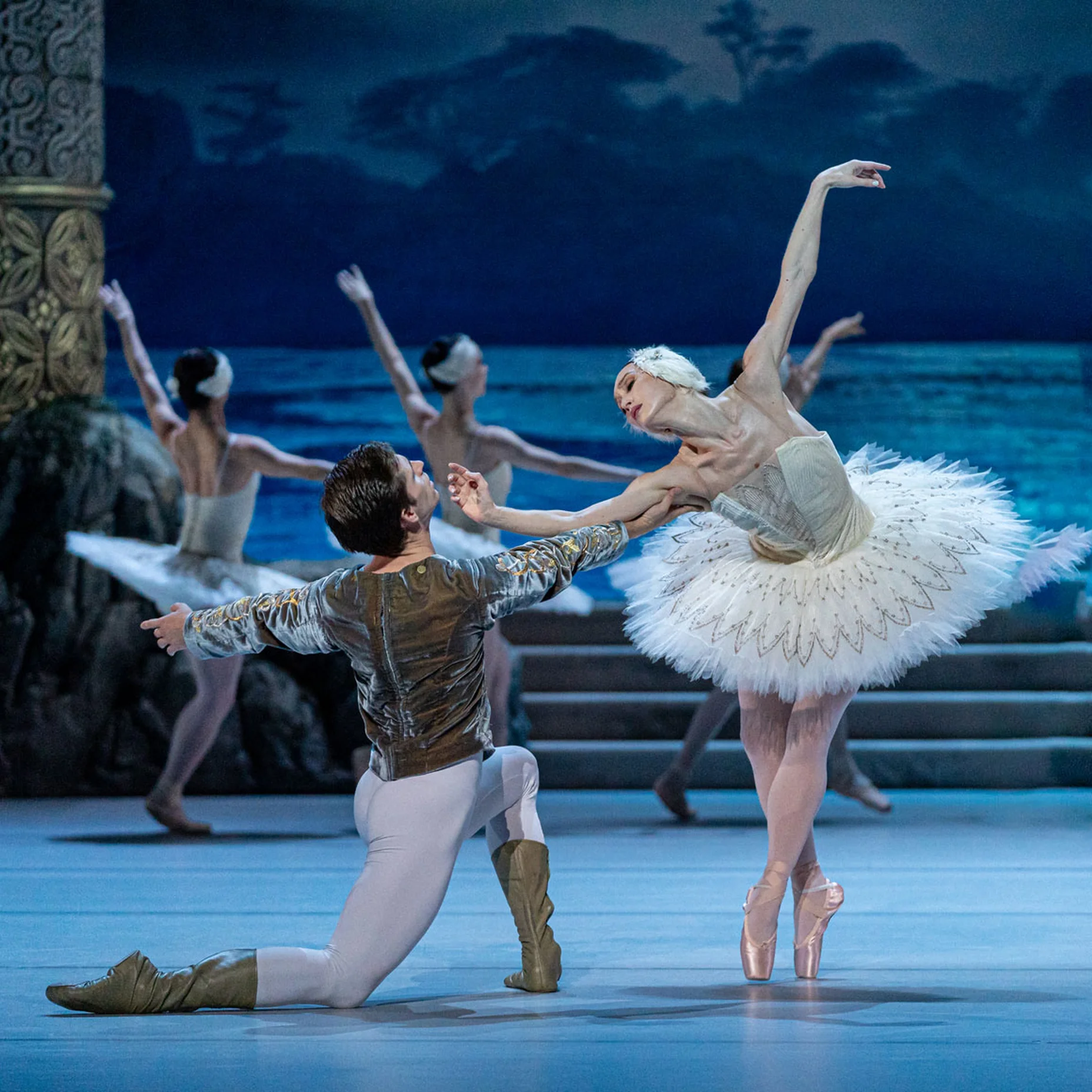 Balletttänzer in klassischer Kleidung führen eine Szene am Ufer eines Sees auf, während Tänzer im Hintergrund zu sehen sind.