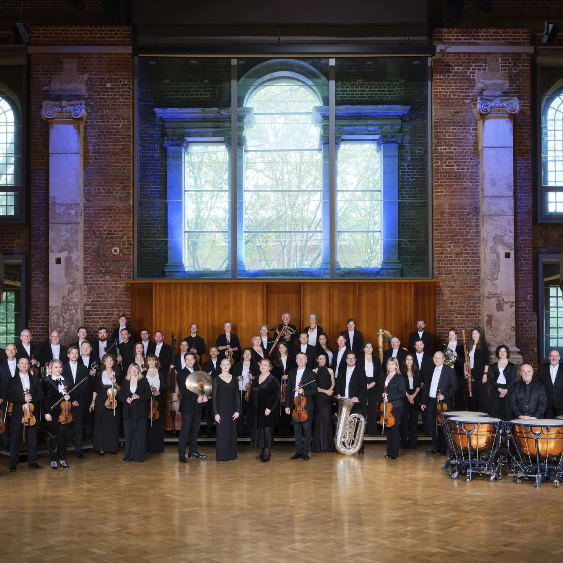 Das London Symphony Orchestra posiert in einer großen Halle mit hohen Fenstern. Musiker halten ihre Instrumente und tragen formelle Kleidung.