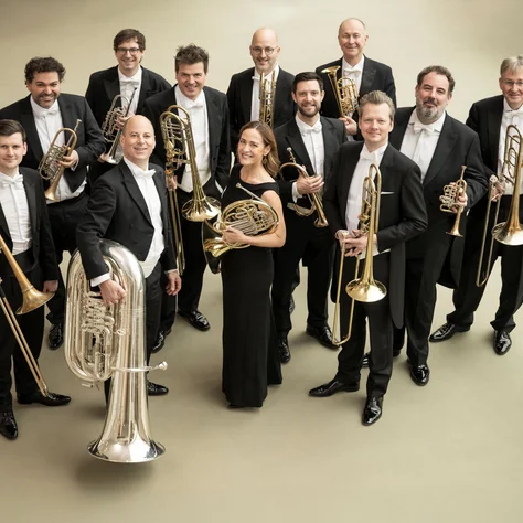 Eine Gruppe von elf Musikern in formeller Kleidung hält Blechblasinstrumente, bereit für eine Aufführung. Sie lächeln in die Kamera.