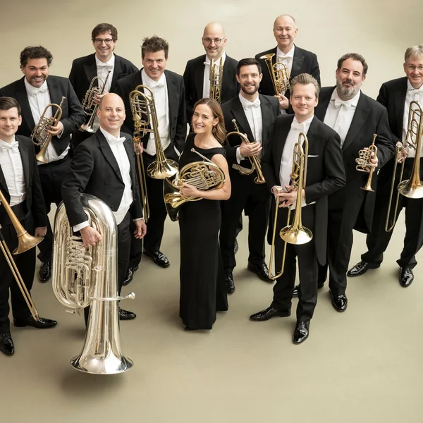Eine Gruppe von elf Musikern in formeller Kleidung hält Blechblasinstrumente, bereit für eine Aufführung. Sie lächeln in die Kamera.
