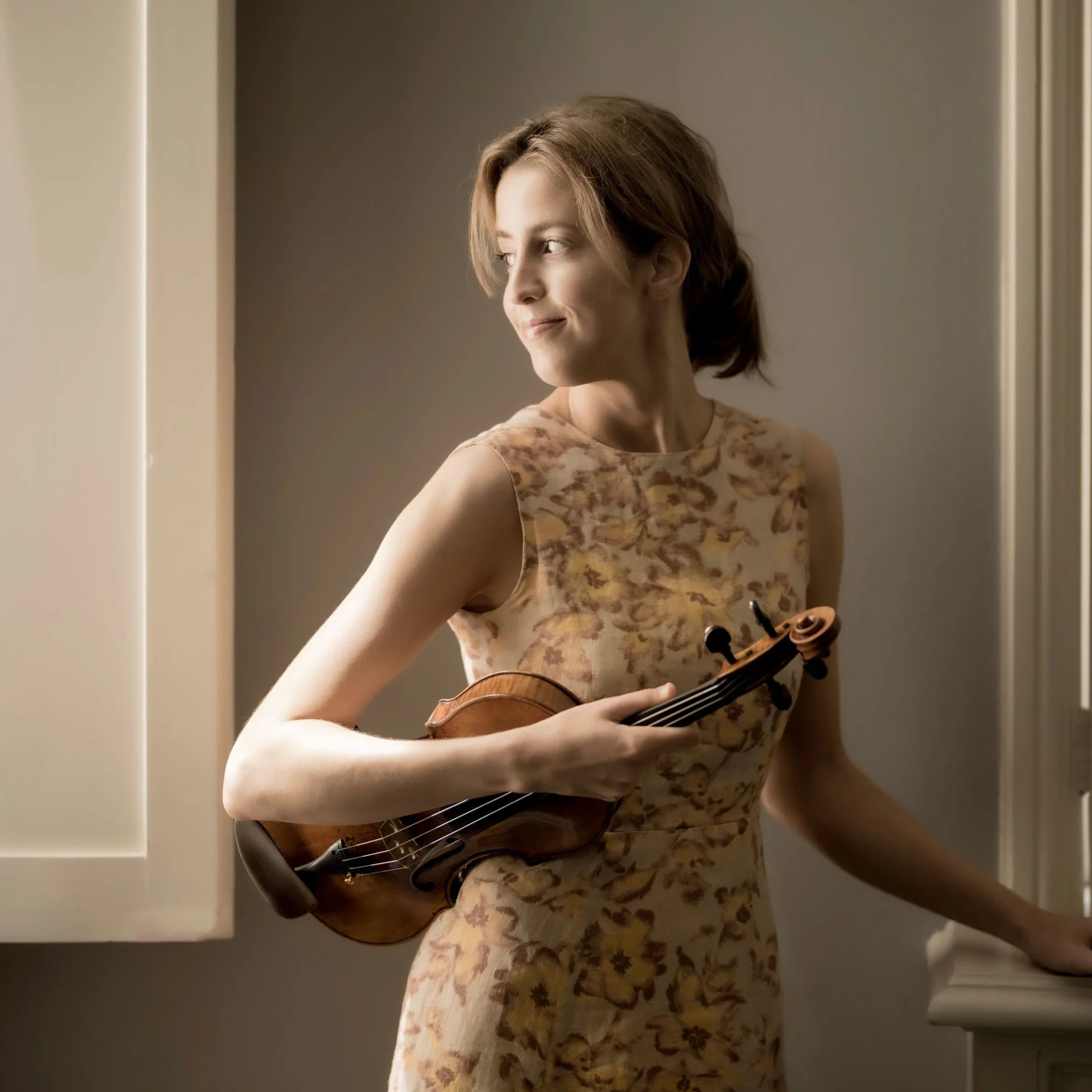 Vilde-Frang in einem eleganten floralen Kleid hält eine Violine und blickt nachdenklich aus einem Fenster.