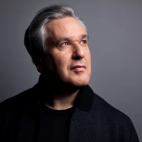 Antonio Pappano with gray hair looks thoughtfully to the side. He wears a dark sweater against a gray background.