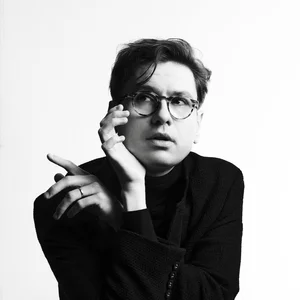 Víkingur Ólafsson mit Brille sitzt an einem Tisch, hält eine Zigarette und schaut nachdenklich zur Seite.