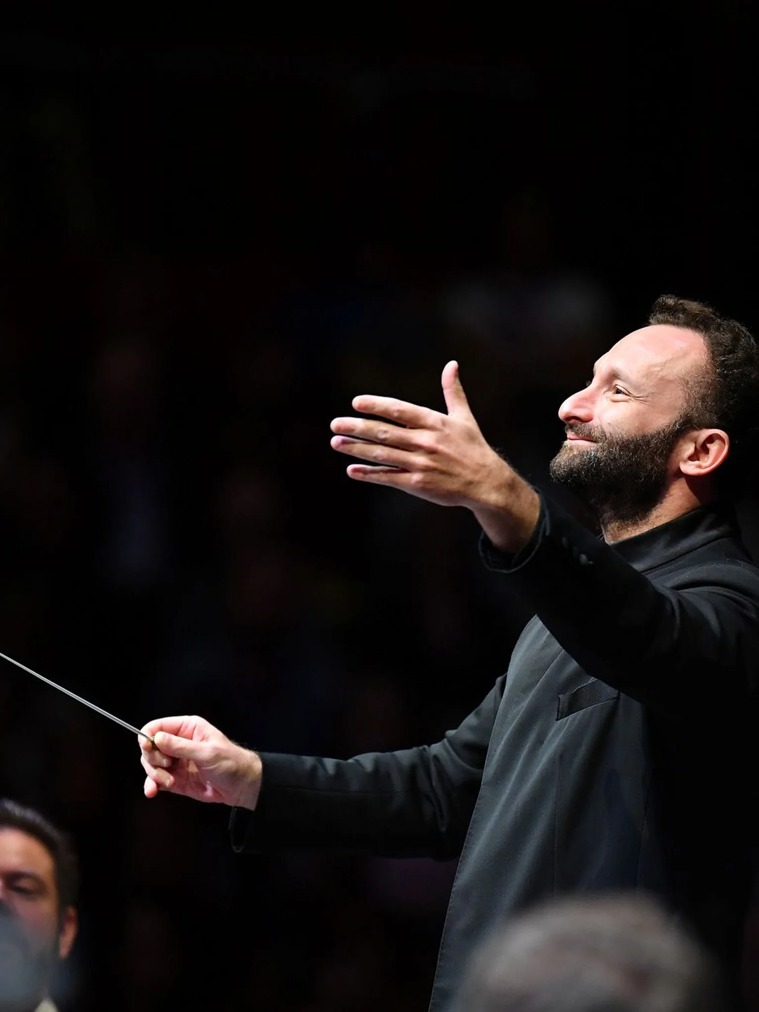 Kirill Petrenko in schwarzer Kleidung leitet mit einem Taktstock ein Orchester. Sein Gesichtsausdruck ist konzentriert und leidenschaftlich.