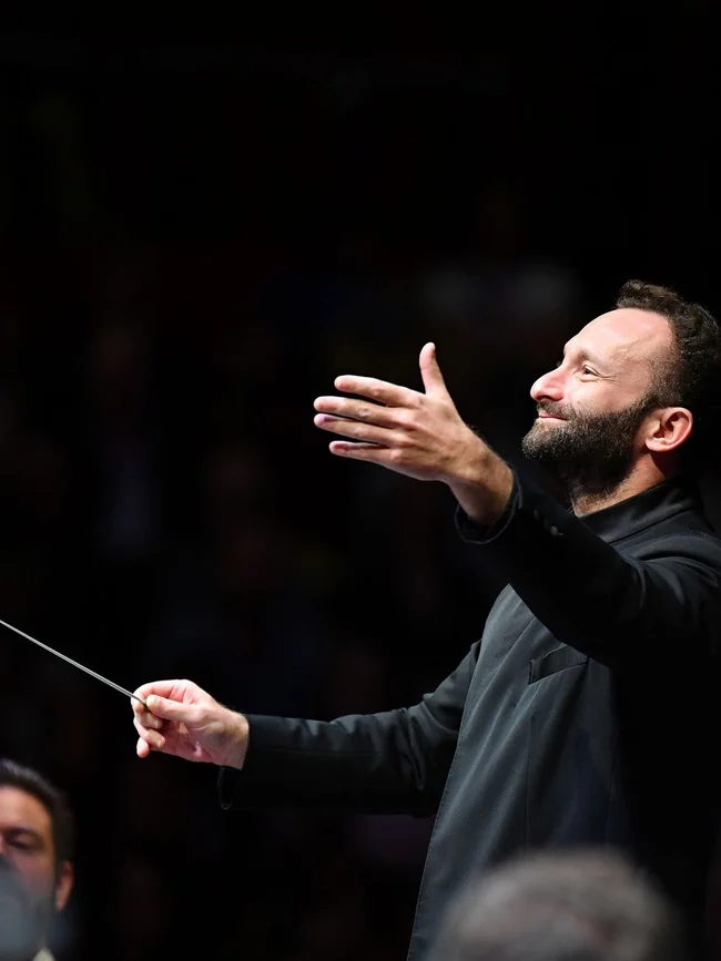 Kirill Petrenko in schwarzer Kleidung leitet mit einem Taktstock ein Orchester. Sein Gesichtsausdruck ist konzentriert und leidenschaftlich.