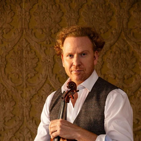 Daniel Hope mit lockigem Haar hält eine Geige und sitzt vor einer dekorativen Wand. Er trägt ein weißes Hemd und eine Weste.
