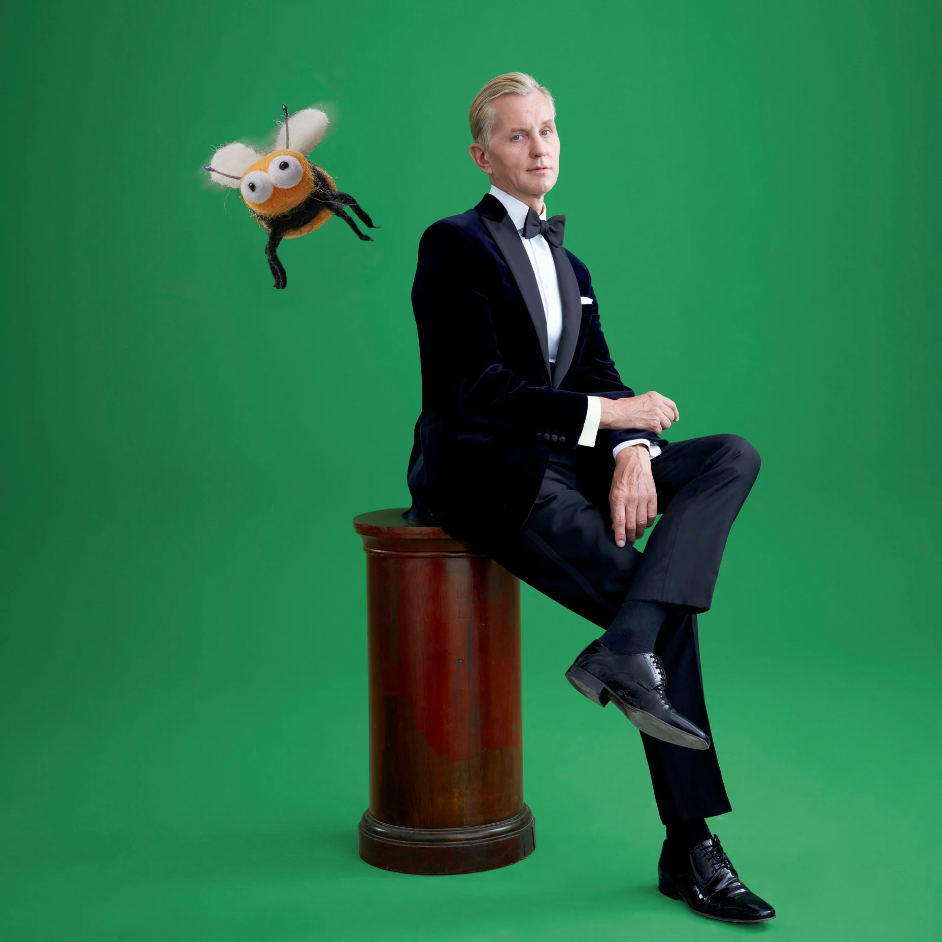 Max Raabe en costume est assis sur un piédestal devant un fond vert. Une abeille dessinée vole à côté.