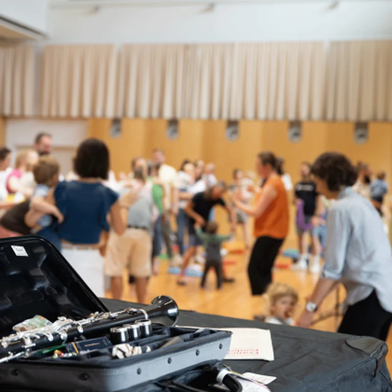 Ein Musiker hat eine Klarinette auf einem Tisch in einem Koffer, während im Hintergrund Leute an einem Krabbelkonzert teilnehmen.