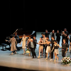 Eine Gruppe von Tänzern steht auf einer Bühne, Menschen applaudieren im Hintergrund. Ein Klavier ist links im Bild.