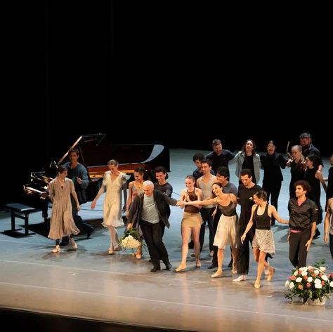 Eine Gruppe von Tänzern steht auf einer Bühne, Menschen applaudieren im Hintergrund. Ein Klavier ist links im Bild.