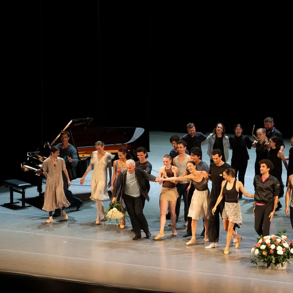 Eine Gruppe von Tänzern steht auf einer Bühne, Menschen applaudieren im Hintergrund. Ein Klavier ist links im Bild.