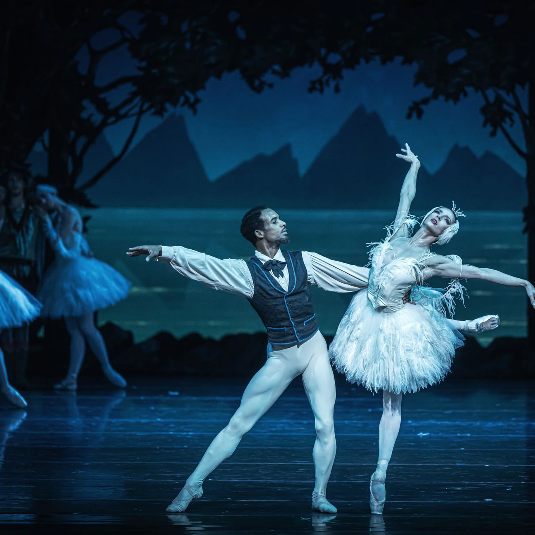 Ein elegantes Ballettpaar performt auf der Bühne. Sie tragen traditionelle Kostüme, mit einem romantischen Hintergrund von Bäumen und Wasser.