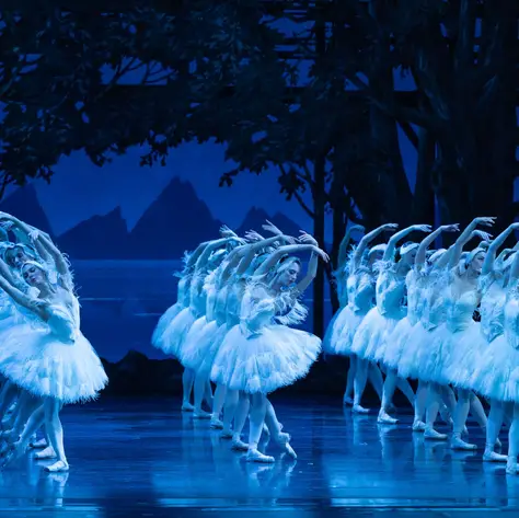 Eine Ballettgruppe in weißen Tutus tanzt synchron auf einer blauen, beleuchteten Bühne, umgeben von einer Waldkulisse. Eleganz und Präzision.