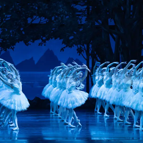 Eine Ballettgruppe in weißen Tutus tanzt synchron auf einer blauen, beleuchteten Bühne, umgeben von einer Waldkulisse. Eleganz und Präzision.