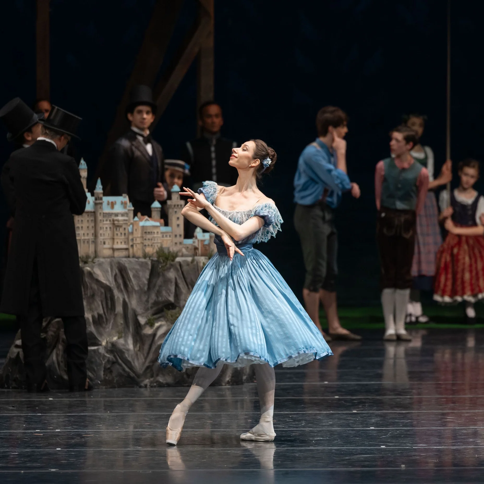 A ballerina in a blue dress performs on stage. In the background, several people dressed in period costumes are visible.