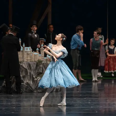 Une ballerine en robe bleue danse sur scène. En arrière-plan, plusieurs personnes en costumes d'époque sont visibles.