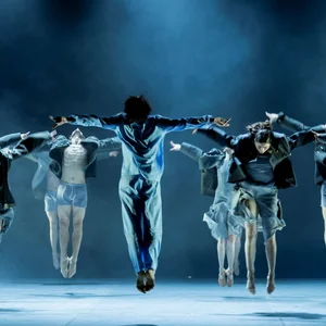 Tänzer des Bundesjugendballetts springen synchron auf einer Bühne, in dramatischem Licht. Die Bewegungen wirken fließend und kraftvoll.