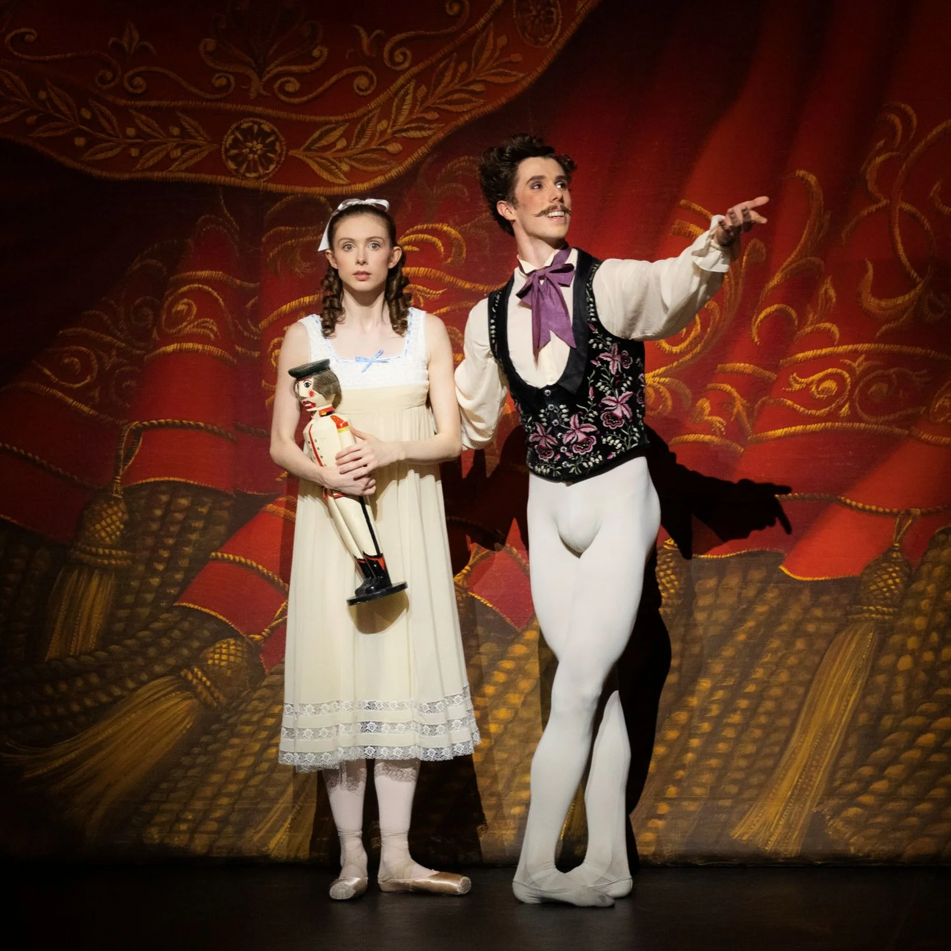 Two dancers in stage costumes stand before an ornate red curtain. One holds a trophy, the other gestures gracefully.