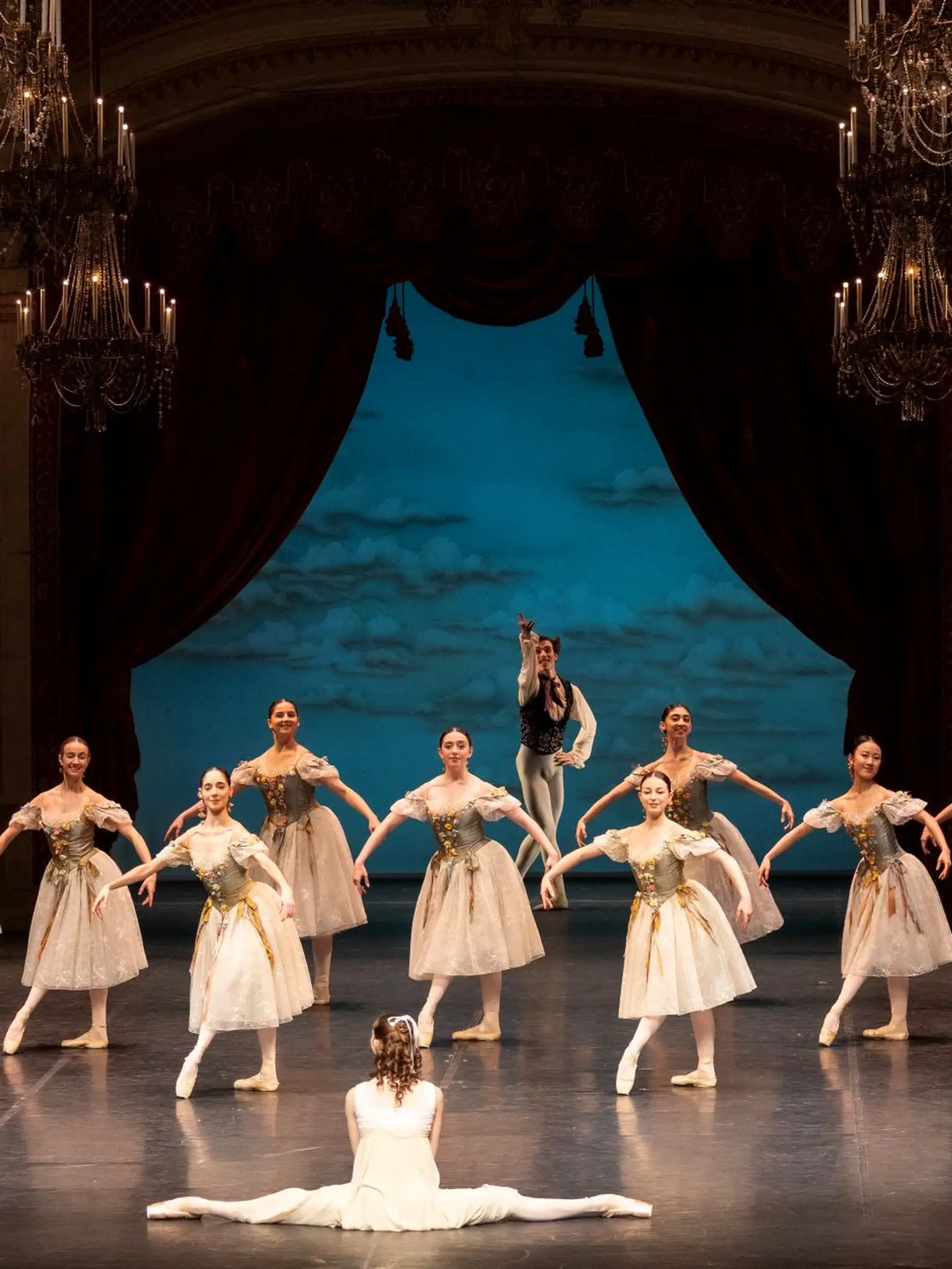 Ballettaufführung auf einer Bühne mit elegant gekleideten Tänzern in weißen Tutus. Eine Tänzerin sitzt im Vordergrund. Eindrucksvolle Kulisse.