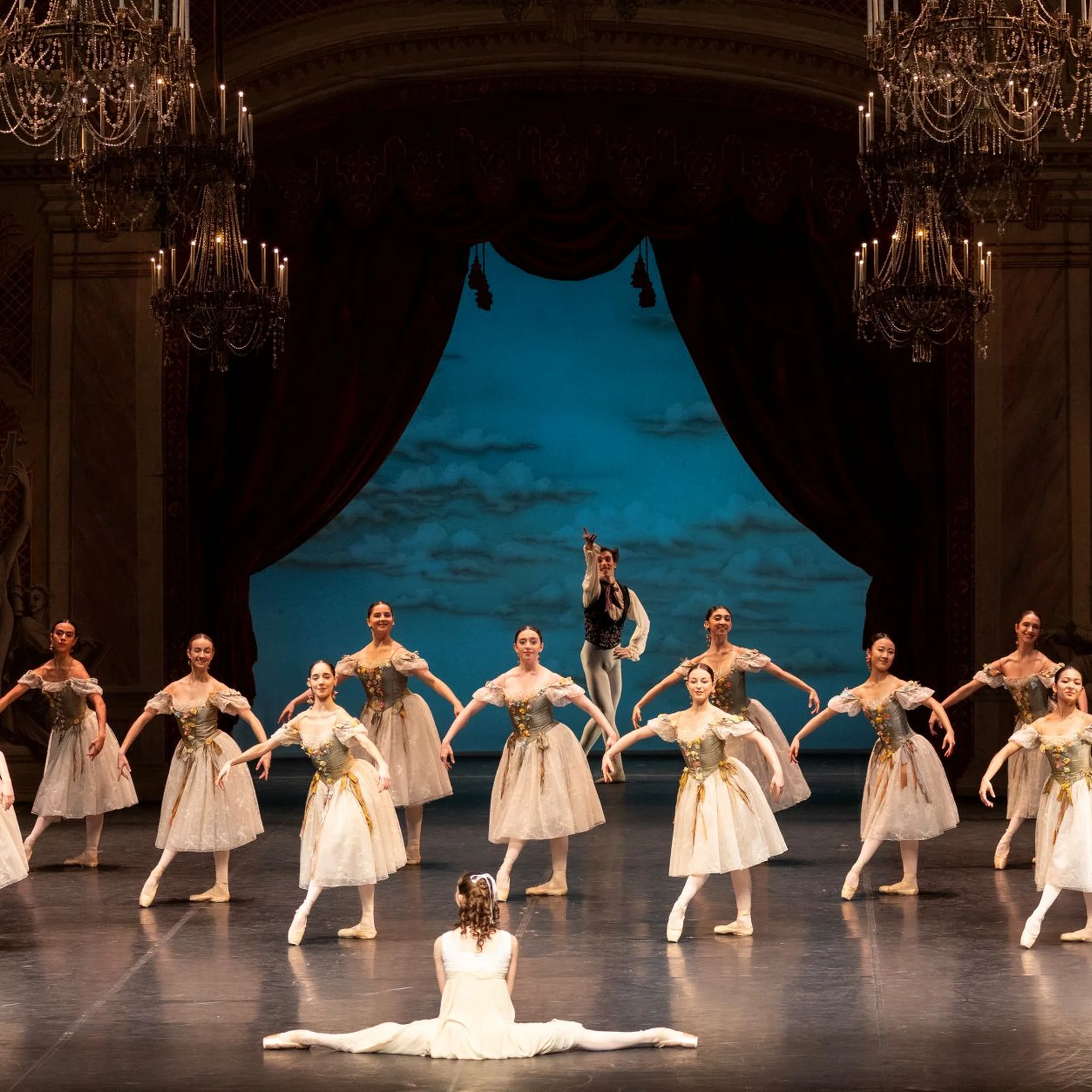 Ballettaufführung auf einer Bühne mit elegant gekleideten Tänzern in weißen Tutus. Eine Tänzerin sitzt im Vordergrund. Eindrucksvolle Kulisse.
