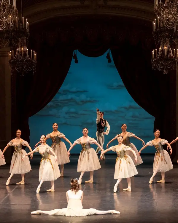 Ballettaufführung auf einer Bühne mit elegant gekleideten Tänzern in weißen Tutus. Eine Tänzerin sitzt im Vordergrund. Eindrucksvolle Kulisse.