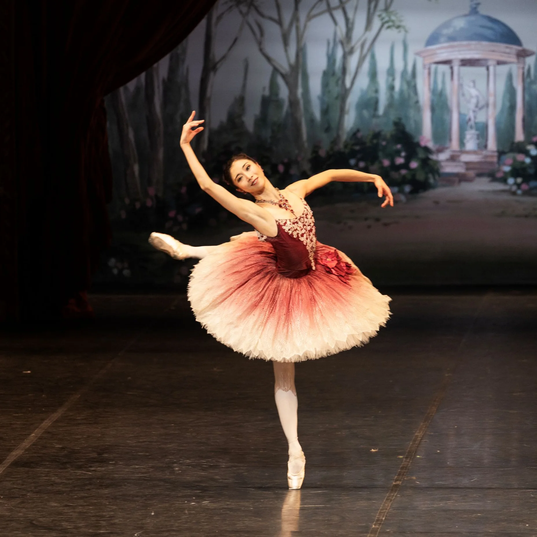 Eine Ballerina führt eine elegante Pose auf der Bühne auf, mit einem pastellfarbenen Bühnenbild im Hintergrund.