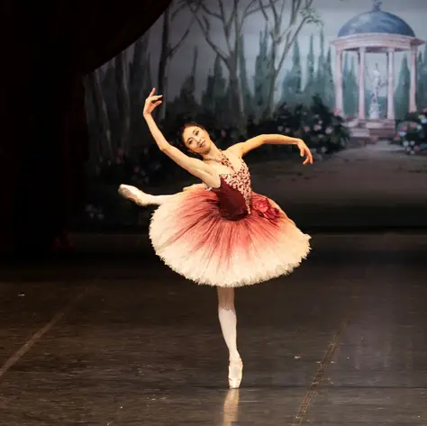 A ballerina performs an elegant pose on stage, with a pastel-colored backdrop in the background.