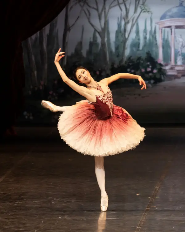 Eine Ballerina führt eine elegante Pose auf der Bühne auf, mit einem pastellfarbenen Bühnenbild im Hintergrund.