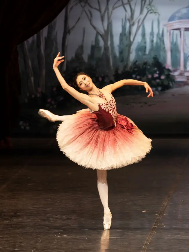 Eine Ballerina führt eine elegante Pose auf der Bühne auf, mit einem pastellfarbenen Bühnenbild im Hintergrund.