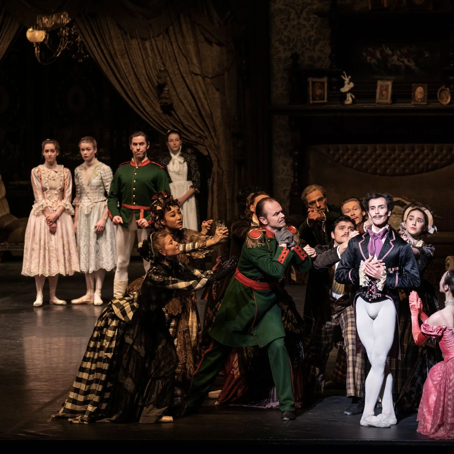 Eine Theaterszene mit mehreren Darstellern in viktorianischen Kostümen. Einige tanzen, während andere im Hintergrund stehen und zuschauen.