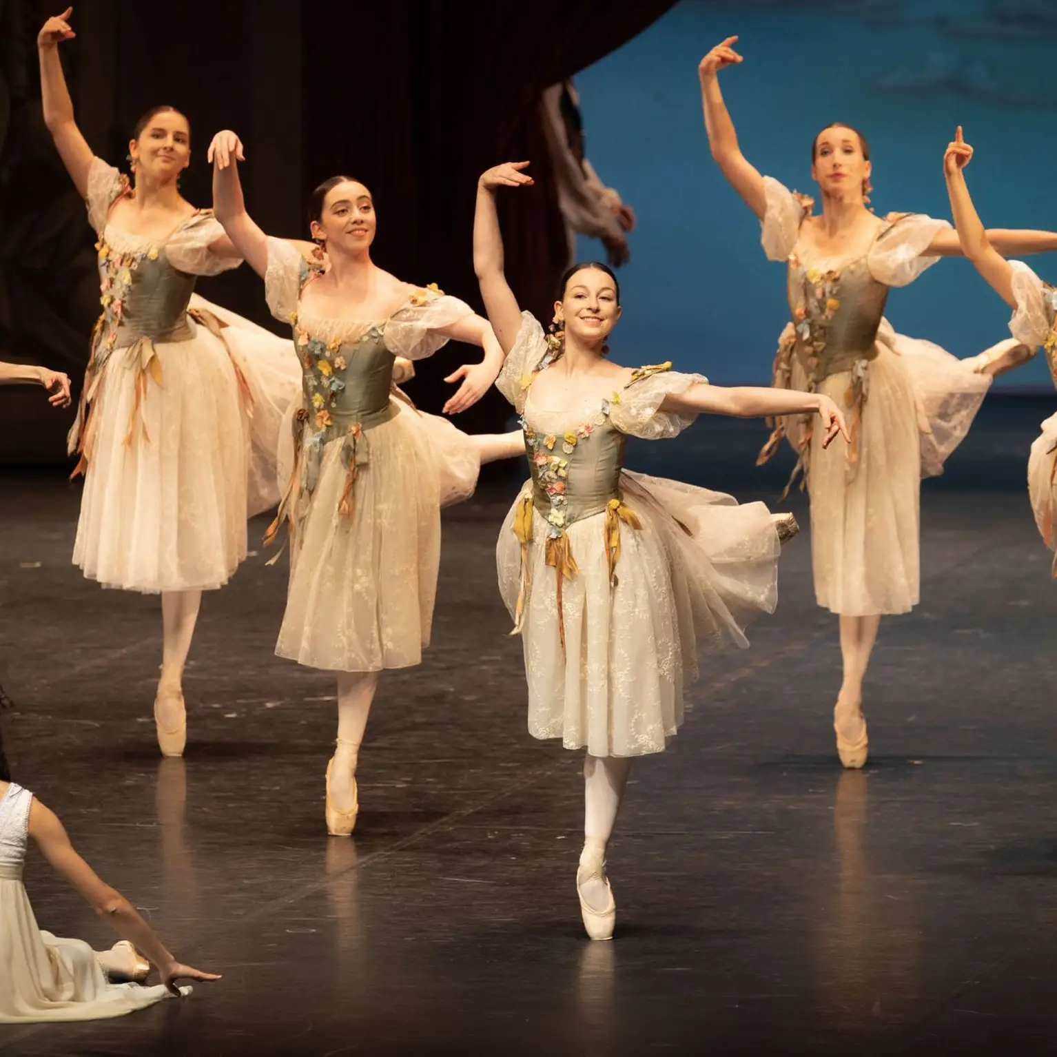 Danseuses de ballet en costumes crème exécutent une performance synchronisée sur scène. Une danseuse est assise à l'avant, observant.