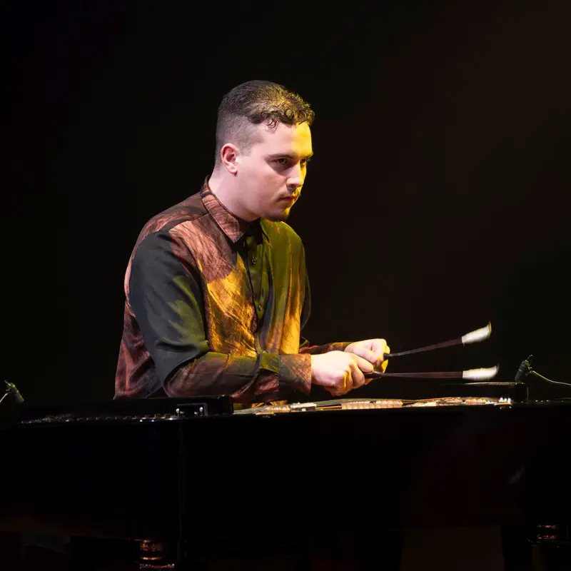 Áron Horváth spielt konzentriert auf einem Vibraphon auf einer stimmungsvoll beleuchteten Bühne. Er trägt ein gestreiftes Hemd.