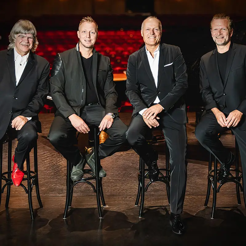 Sebastian Knauer, Martin Tingvall, Joja Wendt und Axel Zwingenberger in schwarzen Anzügen sitzen auf Barhockern auf einer Bühne. Ein Klavier ist im Hintergrund zu sehen.