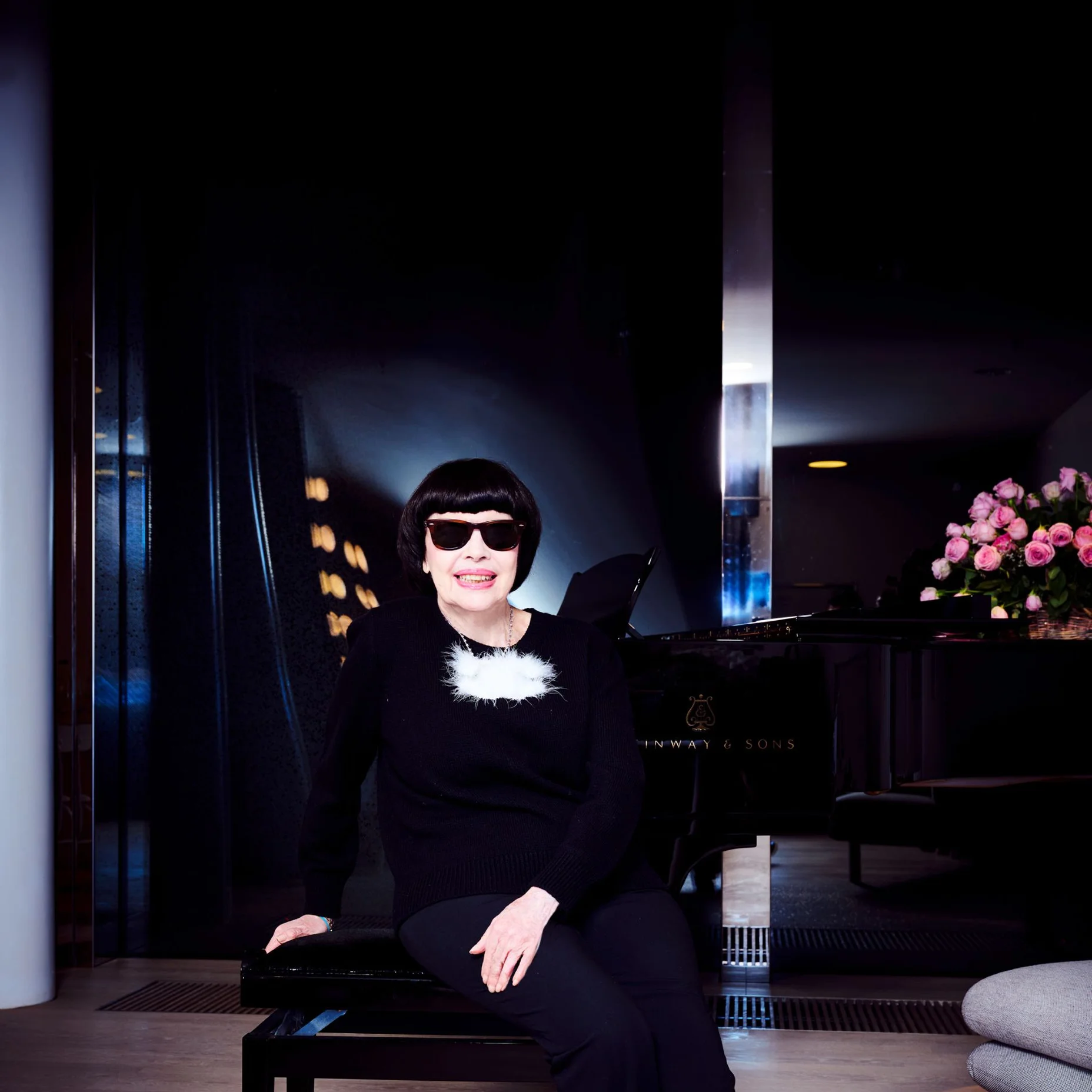 Mireille Mathieu est assise sur une chaise dans une pièce élégante avec un piano en arrière-plan. Elle porte une tenue noire.