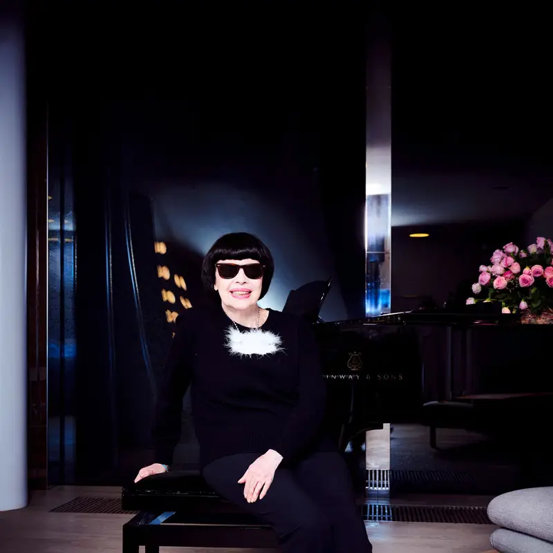 Mireille Mathieu sitzt auf einem Stuhl in einem eleganten Raum mit einem Klavier im Hintergrund. Ihr Outfit ist schwarz.