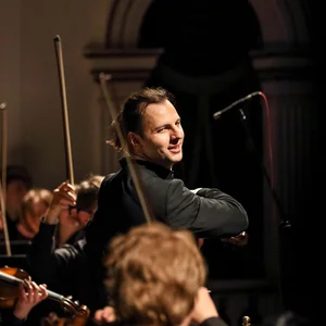 Teodor Currentzis leitet ein Orchester während eines Konzerts. Im Hintergrund sieht man Musiker mit Streichinstrumenten. Die Atmosphäre wirkt konzentriert.
