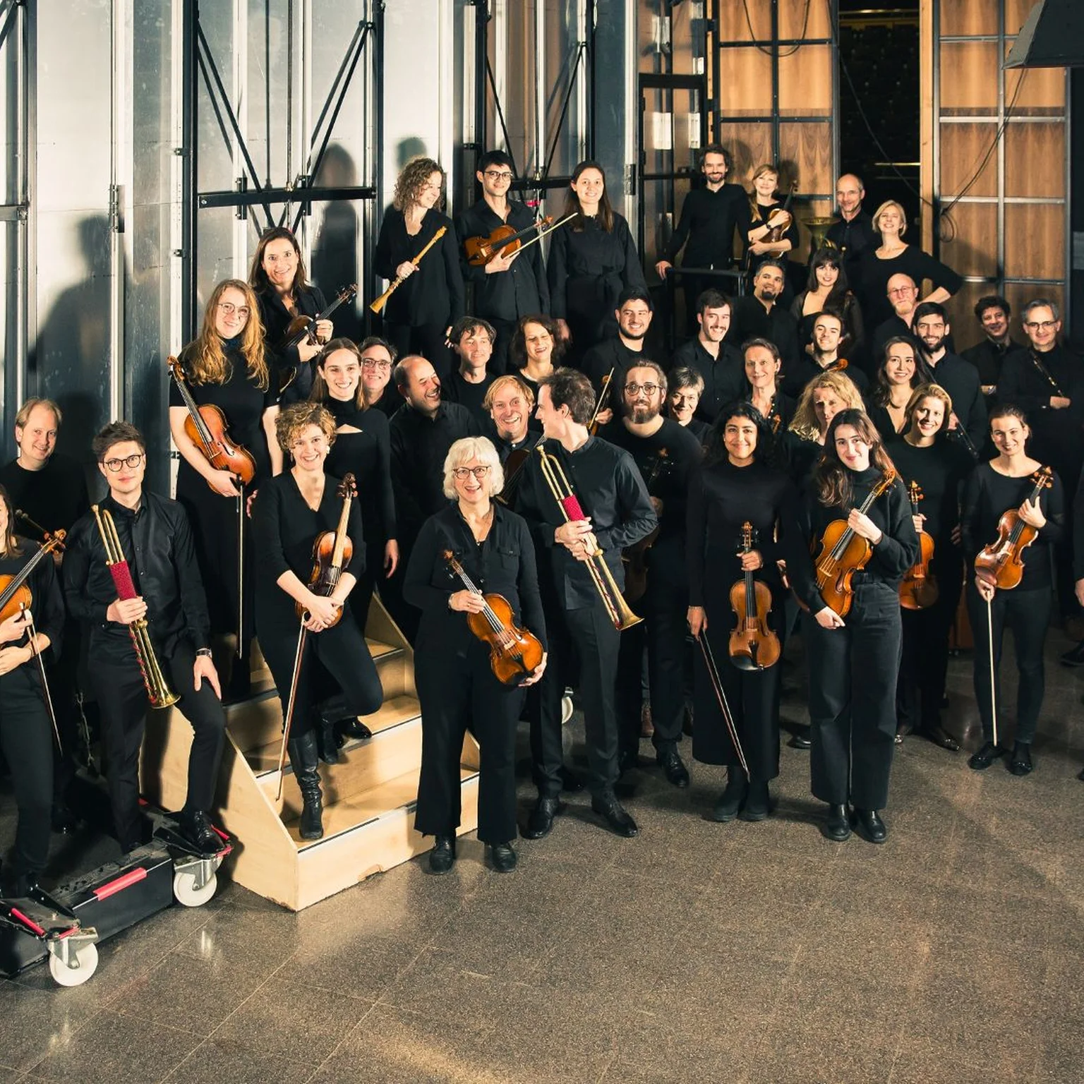 Das Balthasar-Neumann-Orchester in dunkler Kleidung steht in einem Industrieraum mit Instrumenten. Sie bereiten sich auf einen Auftritt vor.