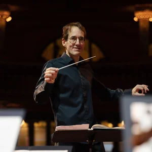 Dirigent Pablo González in Aktion leitet ein Orchester, im Hintergrund sind verschwommene Lichter eines Konzertsaals zu sehen.