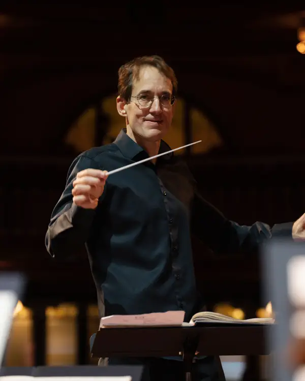Dirigent Pablo González in Aktion leitet ein Orchester, im Hintergrund sind verschwommene Lichter eines Konzertsaals zu sehen.