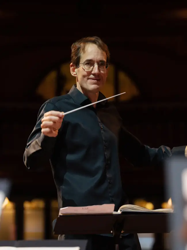 Dirigent Pablo González in Aktion leitet ein Orchester, im Hintergrund sind verschwommene Lichter eines Konzertsaals zu sehen.
