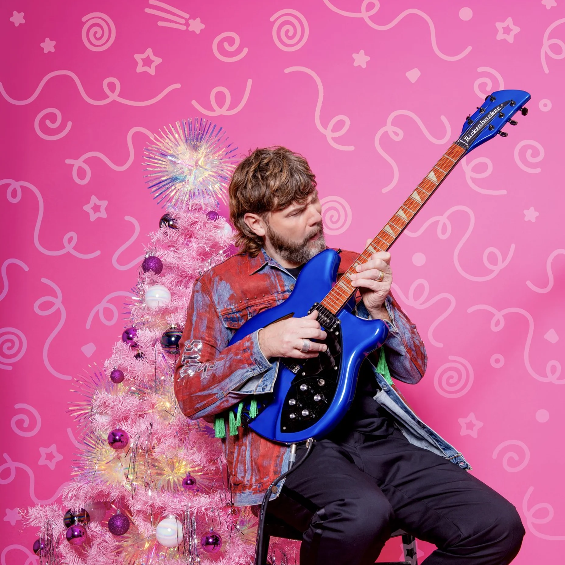 Rea Garvey spielt eine blaue Gitarre vor einem pinken Hintergrund mit Weihnachtsbaum. Die Szene ist bunt und festlich.