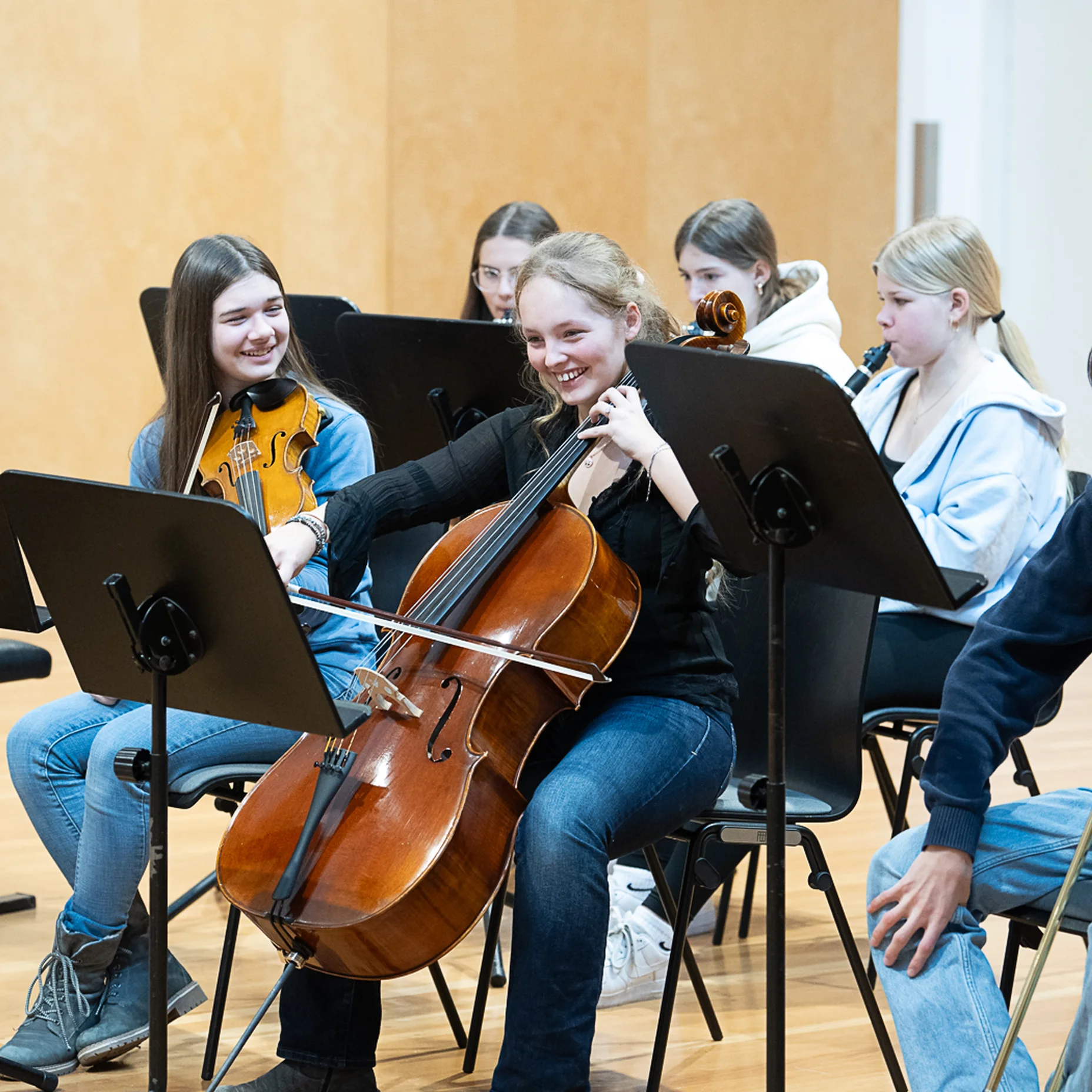Eine Gruppe junger Musiker sitzt in einem Orchesterraum und spielt verschiedene Instrumente, darunter Cello und Trompete.