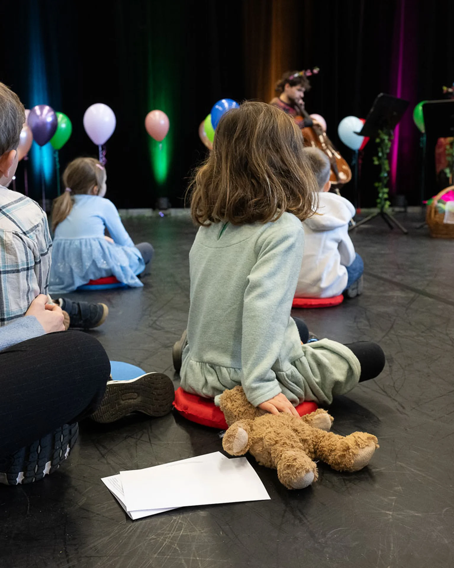 Kinder sitzen im Kreis auf dem Boden, hören Musikern auf einer Bühne mit bunten Ballons zu. Ein Plüschtier liegt neben einem Kind.