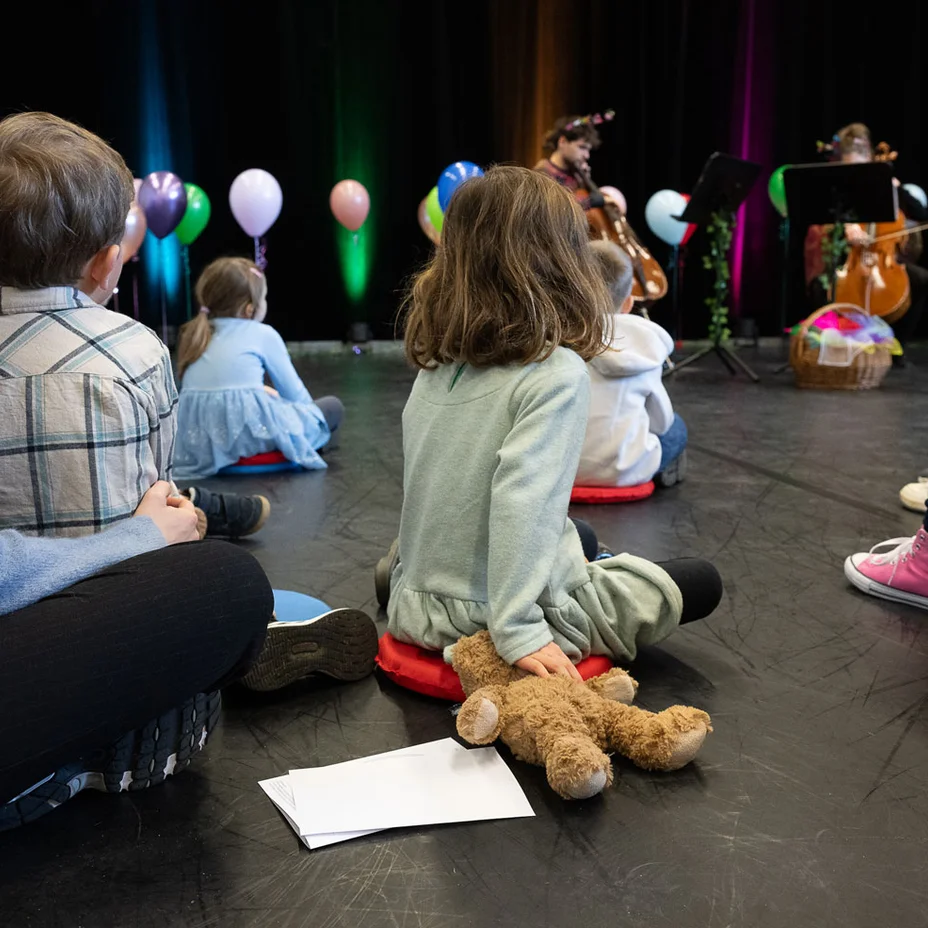 Kinder sitzen im Kreis auf dem Boden, hören Musikern auf einer Bühne mit bunten Ballons zu. Ein Plüschtier liegt neben einem Kind.