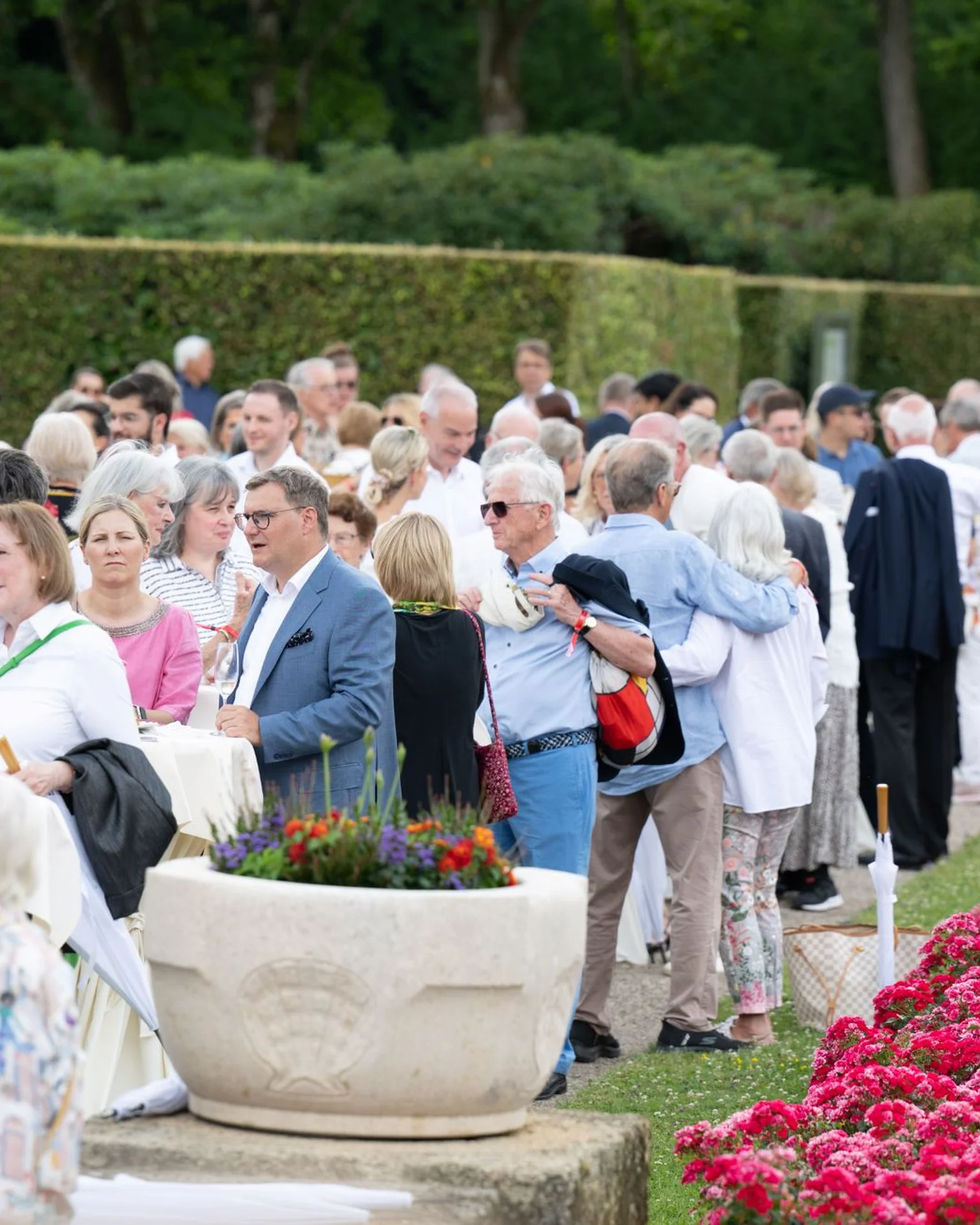 Eine Gruppe elegant gekleideter Menschen versammelt sich in einem gepflegten Garten mit Blumenbeeten und großen Pflanzgefäßen für ein gesellschaftliches Ereignis.
