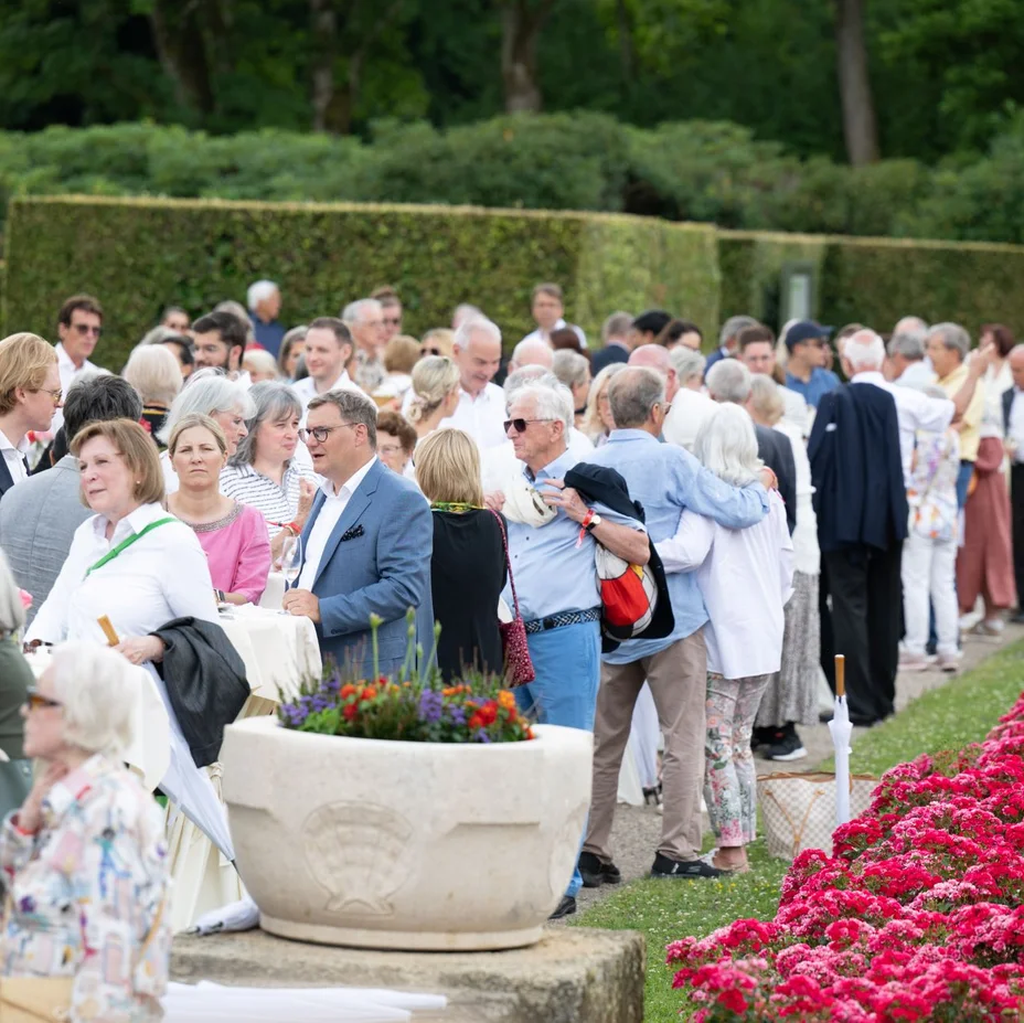 Eine Gruppe elegant gekleideter Menschen versammelt sich in einem gepflegten Garten mit Blumenbeeten und großen Pflanzgefäßen für ein gesellschaftliches Ereignis.